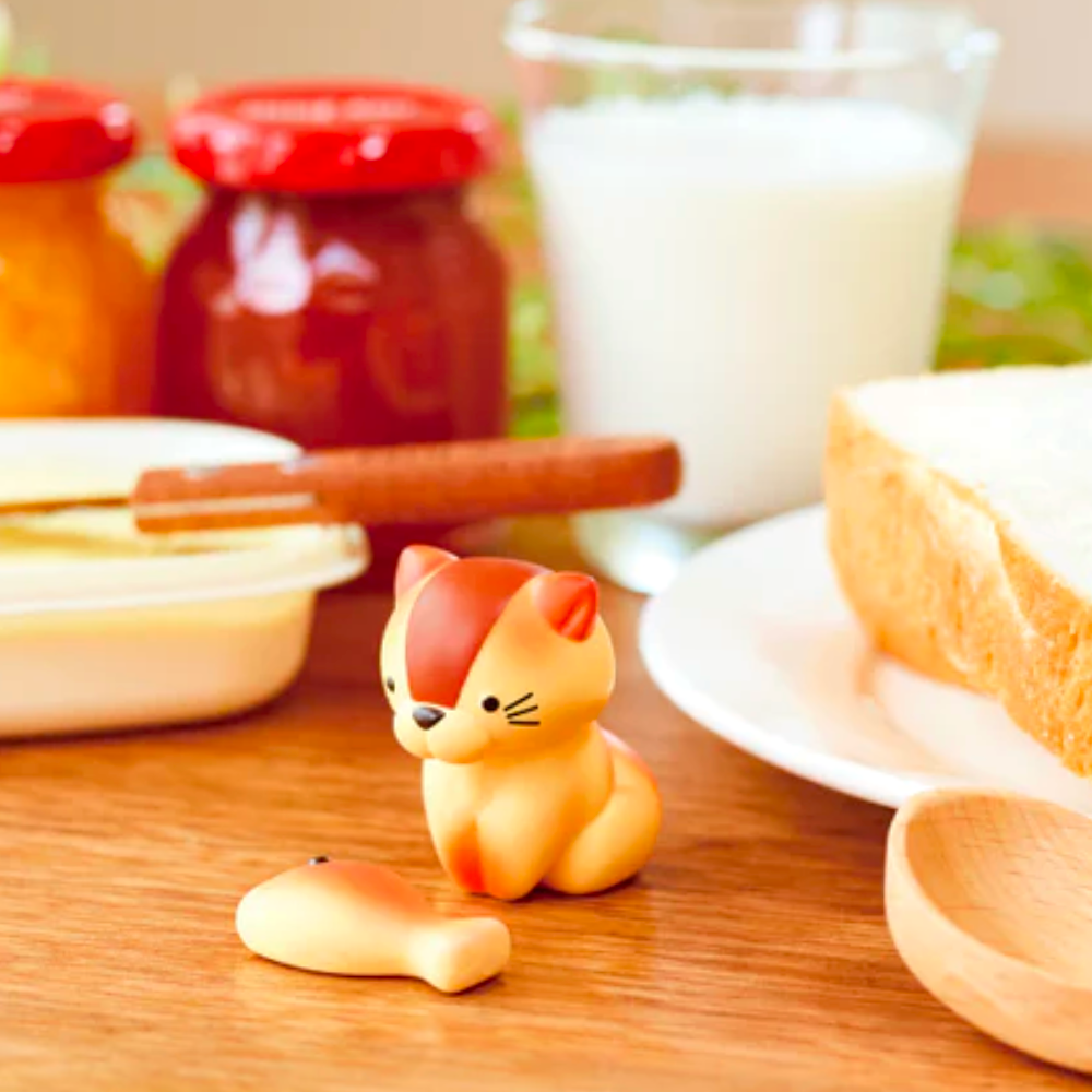 Close-up of an Animal Bakery Blind Box figurine by Dreams and a fish-shaped object on a table with rabbit-shaped bread, a glass of milk, butter dish, and two jars with red lids in the background.