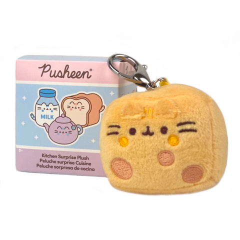 A clip-on plush keychain from Gund's "Pusheen — Kitchen Surprise Blind Box" featuring a bread loaf plush toy with a Pusheen cat design. In the background, there's a small pastel blue and purple box adorned with illustrations of kitchen items and labeled "Pusheen Kitchen Surprise Plush.