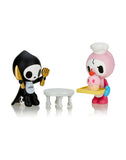 Two stylized figurines: one dressed as a reaper holding utensils, seated at a white table; the other in a chef's outfit holding a tray with a cupcake topped with a cherry. Part of the enchanting Tokidoki Adios & Ciao Ciao Together Forever Blind Box series from tokidoki, they celebrate the love between life and death in culinary art.