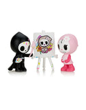 Two cartoon skeleton figures, one in black painting clothes holding a palette and brush, the other in pink, stand by an easel displaying a painted skeleton artwork. This whimsical scene is part of the 