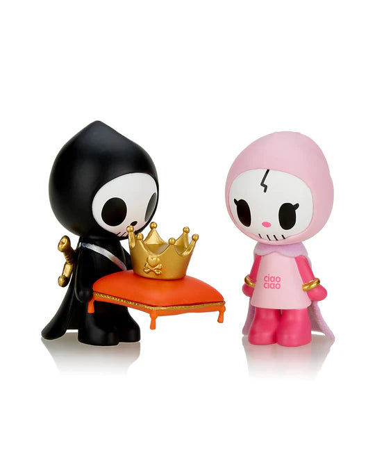 The Tokidoki Adios & Ciao Ciao Together Forever Blind Box by tokidoki features two distinct cartoon figurines: one character donning a black cloak while holding a small golden crown on a red pillow, and the other dressed in pink with a small scar on its forehead, perfectly capturing their quirky love and unique personalities.