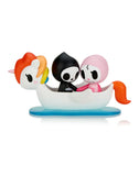 Two small figurines, Adios & Ciao Ciao from the tokidoki brand, featuring skull-like faces—one in black and the other in pink—sit in a white unicorn-shaped boat on a blue base. The Tokidoki Adios & Ciao Ciao Together Forever Blind Box is perfect for fans of the Blind Box Series or anyone looking to add a touch of whimsical love to their collection.