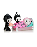 A cartoon figure in black presents a gift to another figure with cat ears, who is sitting beside a sleeping figure in bed with a pink blanket adorned with skull designs, capturing a moment of love from the tokidoki Adios & Ciao Ciao Together Forever Blind Box Series.