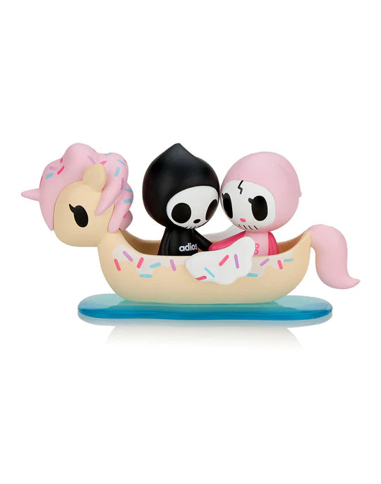 Two small, stylized cartoon characters, Adios & Ciao Ciao, in black and pink outfits ride in a whimsical, unicorn-themed boat with sprinkles on a blue base. This adorable scene is part of the Tokidoki Adios & Ciao Ciao Together Forever Blind Box series by tokidoki, capturing the essence of love and adventure.