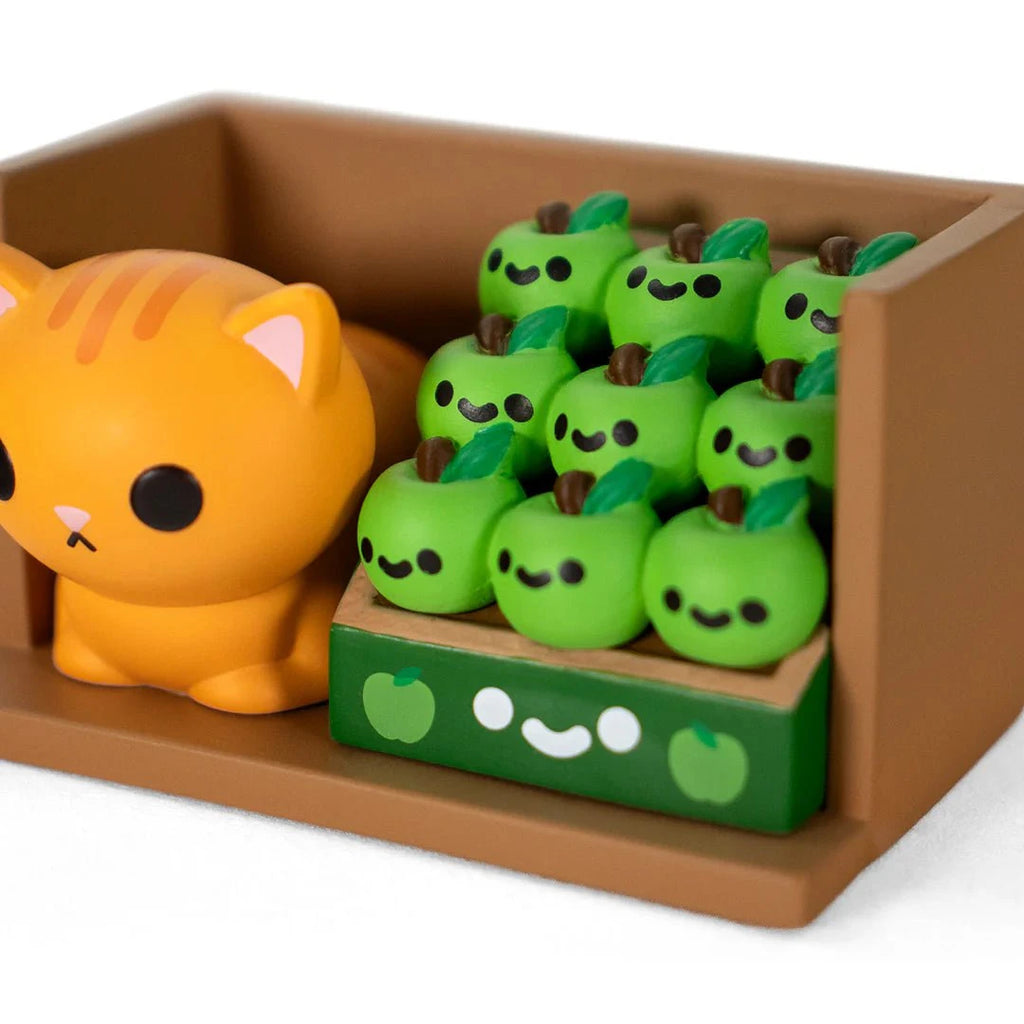 A small orange Bodega Kitties — Series 2 figurine by 100% Soft sits beside a Blind Box display of green smiling apple figures, all arranged on a brown tray.
