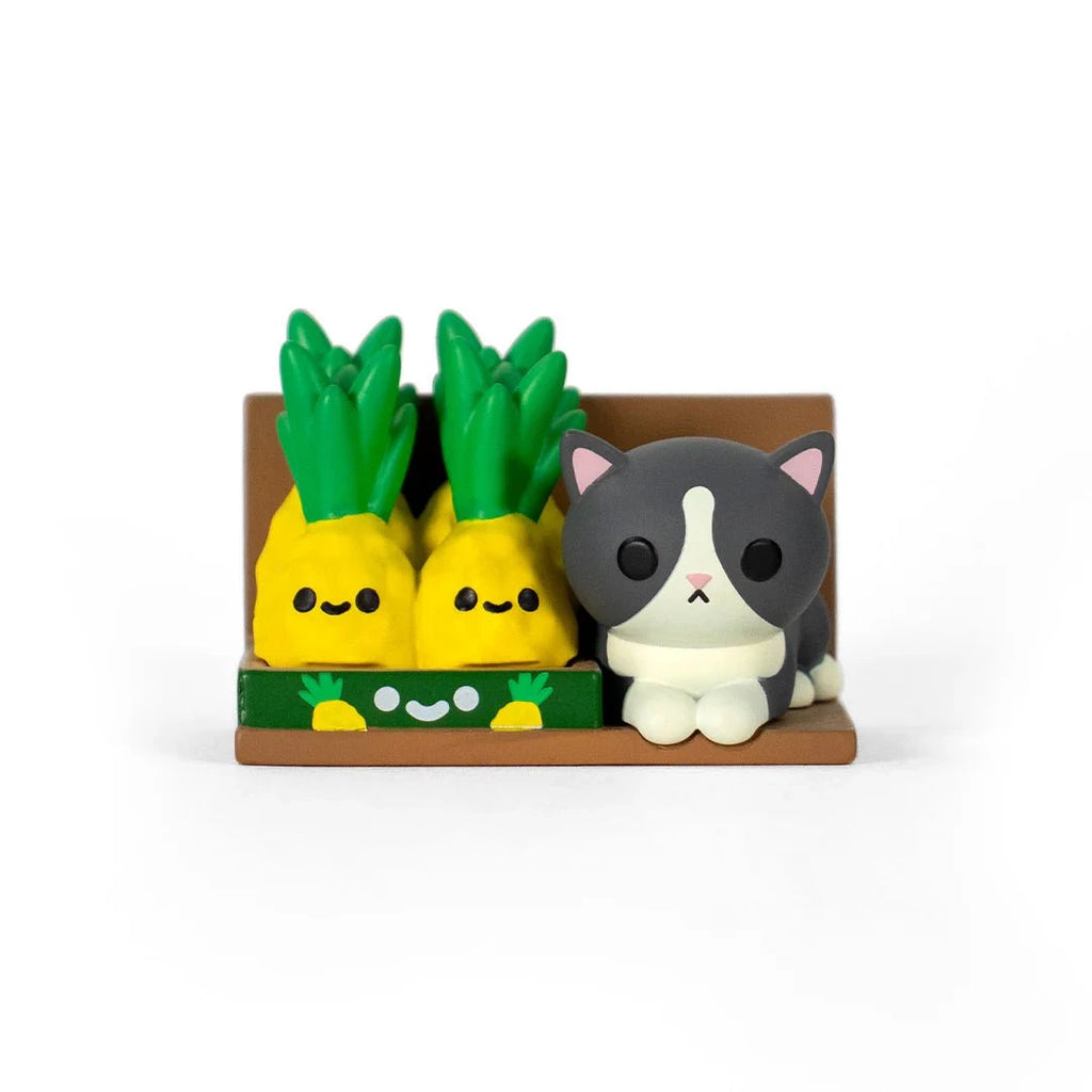 A gray and white cat figurine sits by two smiling pineapples on a brown shelf—just like a surprise from the 100% Soft Bodega Kitties — Series 2 Blind Box.