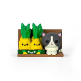 A gray and white cat figurine sits by two smiling pineapples on a brown shelf—just like a surprise from the 100% Soft Bodega Kitties — Series 2 Blind Box.
