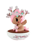 A small pink and brown deer-like figurine with wings and a flower crown from the tokidoki Botanical Bonsai Unicorno Blind Box sits in a white bowl with cherry blossom designs on a brown surface.