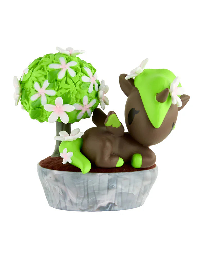 A small brown-and-green deer-like figurine with white flowers rests by a blooming tree in a gray planter—an adorable collectible from the tokidoki Botanical Bonsai Unicorno Blind Box, perfect for miniature garden or tokidoki fans.