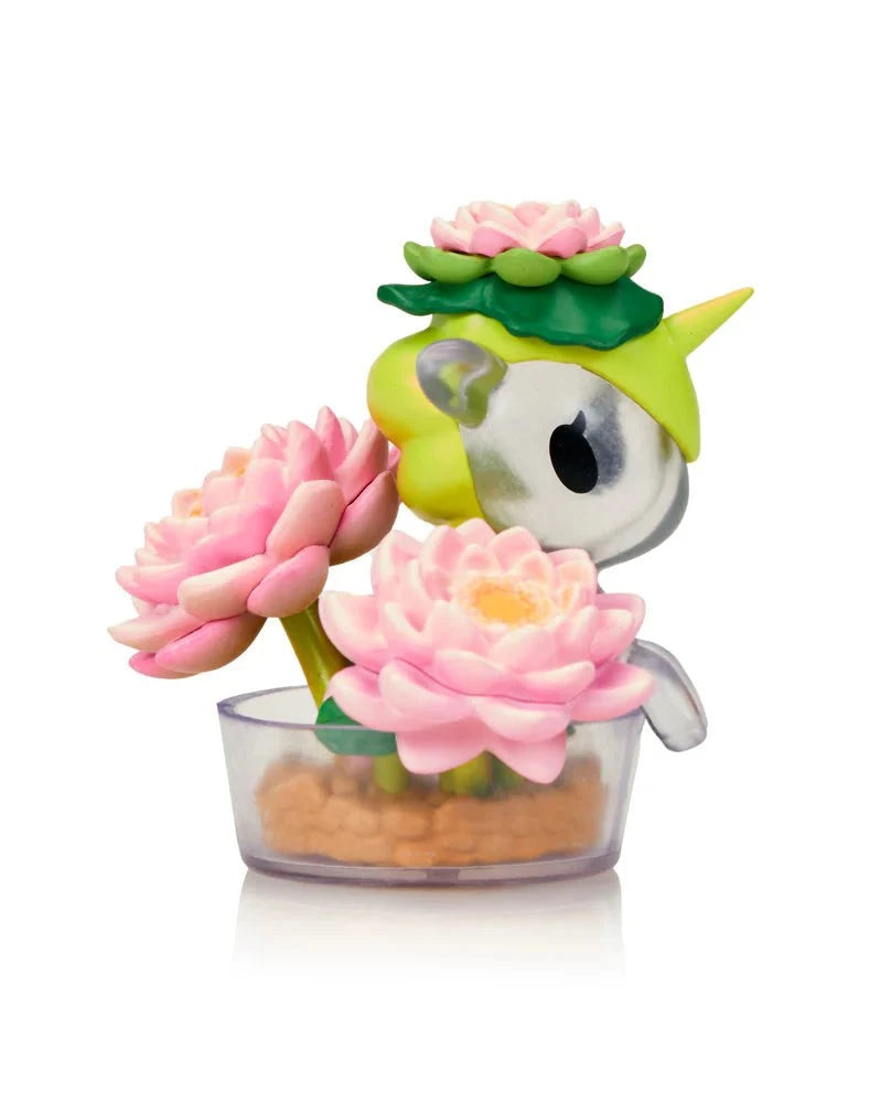 The Tokidoki Botanical Harmony Unicorno Blind Box features a small Tokidoki figurine with green and yellow accents, displayed among pink lotus flowers in a clear container of tan pebbles against a white background.