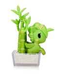 A Tokidoki Botanical Harmony Unicorno Blind Box figurine featuring gold accents sits beside plastic bamboo in a white planter, bringing a natural touch indoors.