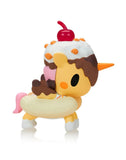 A Tokidoki Ice Cream Shop Unicorno Blind Box toy figure by Tokidoki, featuring a donut-shaped body with chocolate icing, a cherry on top, and a pink tail, is displayed standing on a white surface.