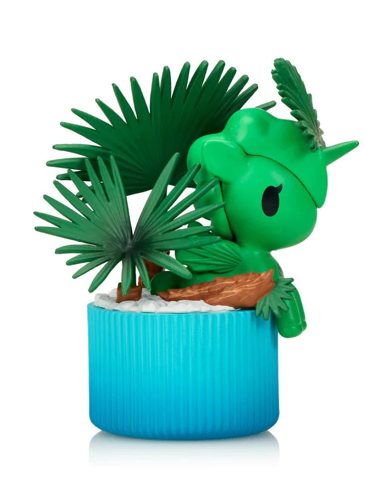 The Tokidoki Botanical Harmony Unicorno Blind Box features a small green figurine with palm leaves on its head and back, sitting on a blue base with white accents. Perfect for collectors, it beautifully captures the essence of nature.
