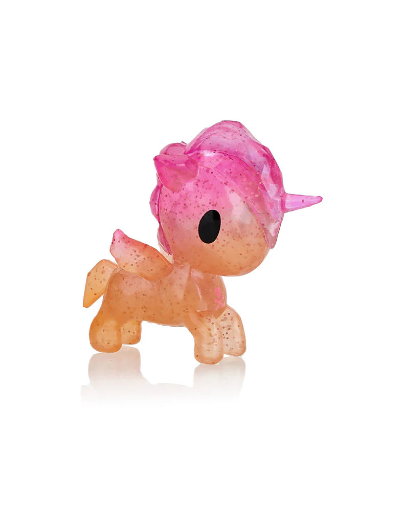 A small, translucent Unicorno Capsule Cuties Pink Dusk Special Edition by Tokidoki, speckled in pink and orange hues with a pink mane and horn, stands elegantly on a reflective surface. This enchanting piece beautifully captures the magical allure of a pink dusk.