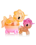 An orange and a pink unicorn figure, both with sparkly textures, are positioned side by side on a white background. These are the Tokidoki Unicorno Capsule Cuties Pink Dusk Special Edition set.