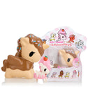 Two figurines from the Tokidoki Toki Mochi Donut Unicorno Blind Bag, including one decorated with sprinkles and a pink one embellished with a strawberry, are displayed prominently in front of their packaging that highlights various enchanting Unicorno characters.