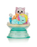 A Tokidoki Dreaming Unicorno Blind Box by tokidoki, featuring a toy figure of a sleeping Unicorno with a colorful mane, accompanied by a small blue seahorse and a cute cat figure, all placed on a transparent green stand, creating the perfect setting for a magical adventure.