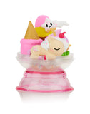 A small figurine of an ice cream-themed Unicorno sleeping in a clear, pink base display case, holding an ice cream cone, with colorful accessories around it. This charming piece captures the essence of a magical adventure. Introducing the Tokidoki Dreaming Unicorno Blind Box by tokidoki.
