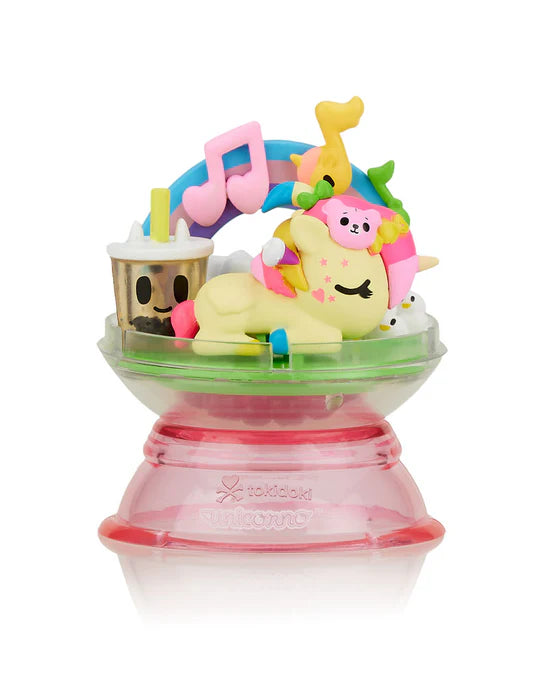 A colorful toy figurine featuring a Dreaming Unicorno with a pink mane lounging on a grassy base, surrounded by a rainbow with music notes and other small characters, all mounted on a pink pedestal. This sets the stage for a magical adventure straight out of the tokidoki Tokidoki Dreaming Unicorno Blind Box collection.
