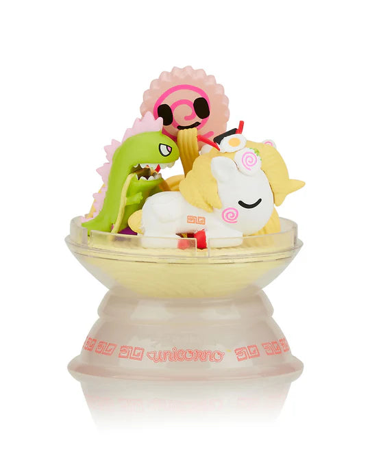 Small figurines including a sleeping unicorn, a green dinosaur, and a pink smiling character are arranged in a bowl-like container with yellow filling. The base reads 
