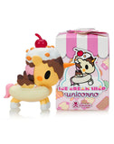 A small unicorn figurine with ice cream and a cherry on its head stands next to the Tokidoki Ice Cream Shop Unicorno Blind Box by Tokidoki, featuring a pink ice cream-themed design.