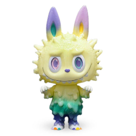 A small toy figure of a whimsical creature with large ears, gradient pastel colors, and a fluffy texture. It has a wide grin, multicolored eyes, and gradient colors from yellow to green to purple is the INC Labubu by How2Work (HK).
