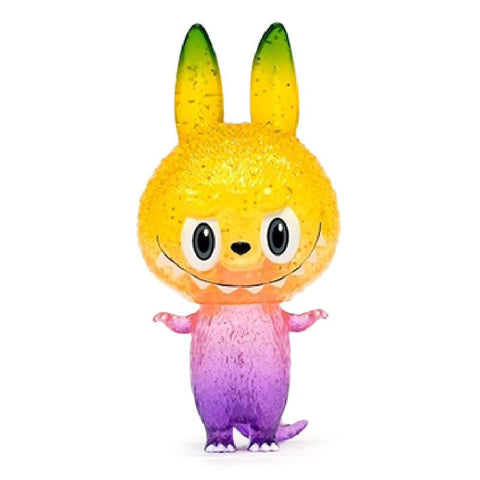 A colorful toy with a large head resembling a cat standing upright. The head is yellow-green and the body is a gradient from pink to purple. The toy has big eyes, pointed ears, and a small smile. This is the Mini Zimomo — Ceibo by How2Work (HK).
