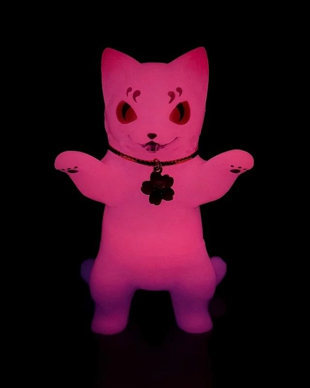The Konatsuya Negora — Night Sakura is a glowing pink, glow-in-the-dark cat figurine with raised paws, a collar, and cherry blossom-shaped tag, shown against a black background.