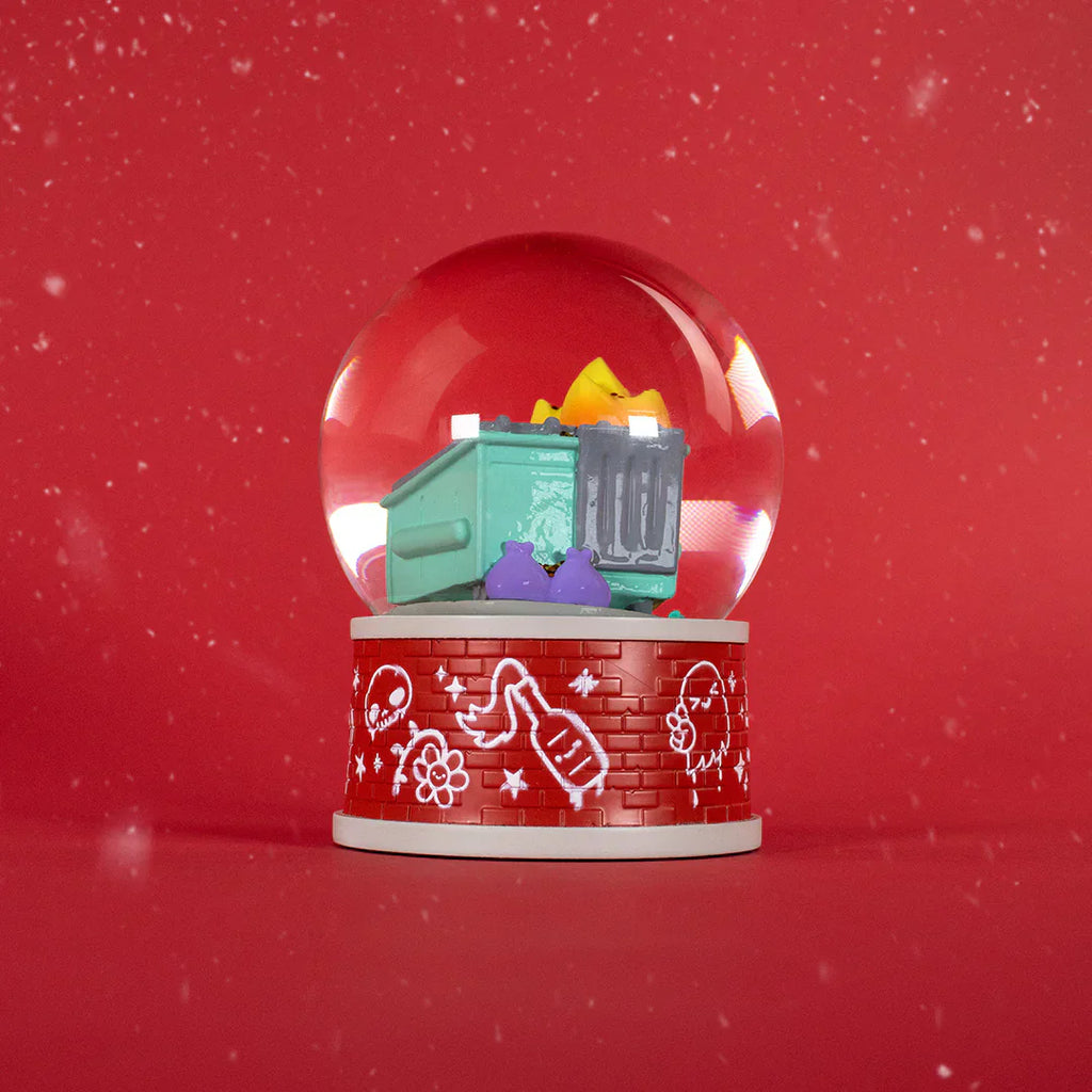 The Dumpster Fire Snow Globe by 100% Soft has a green dumpster with yellow and purple trash on a red base with white line art, making quirky seasonal desk decor against a red background with white specks.