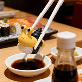 Chopsticks hold a Rotofugi Sushi Snails Blind Box figure shaped like a yellow snail above soy sauce, with additional sushi and condiments in the background.
