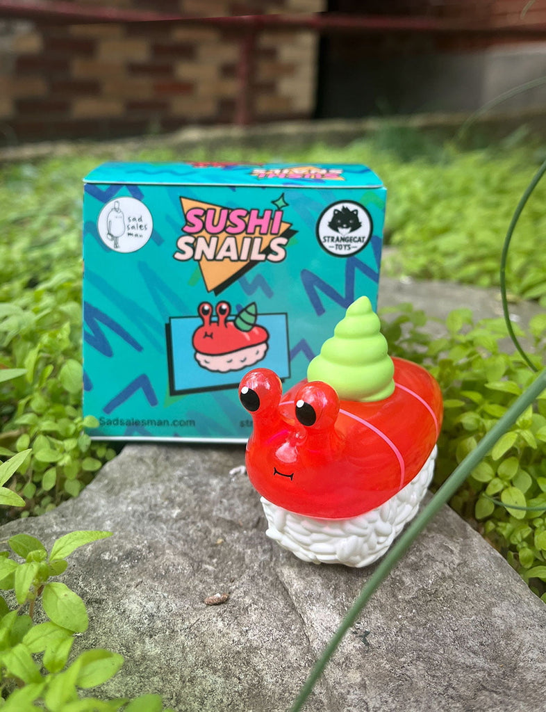 A Rotofugi Sushi Snails Blind Box collectible, featuring a toy snail styled like sushi on a stone with green plants, is shown with its colorful packaging in the background—ideal for fans of fun art toys.