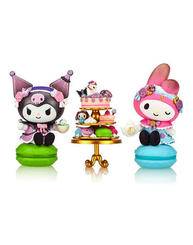 Two cute figurines holding tea cups are displayed on green and blue macarons. Between them is a small, ornate table with cakes and pastries. Both characters, inspired by Kuromi & My Melody, wear colorful outfits and headpieces in this charming Tokidoki tokidoki x Kuromi & My Melody Garden Party - Garden Tea Party (Special Edition 2-pack) scene.