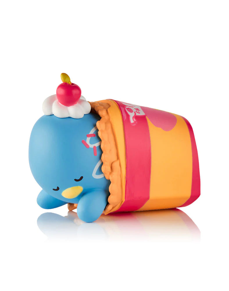 A blue cartoon character with a cherry and whipped cream on its head is wrapped in a crepe-like blanket, capturing the playful style of collectible figures from the tokidoki x Hello Kitty & Friends Fast Food Blind Box by tokidoki (IT).
