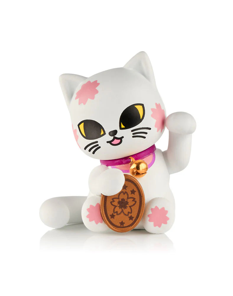 A collectible kitty figurine from the tokidoki (IT) Tokidoki Lucky Cats Blind Box, featuring a waving cat with pink flower patterns, yellow eyes, a purple collar with a bell, and a brown oval coin with a flower design.