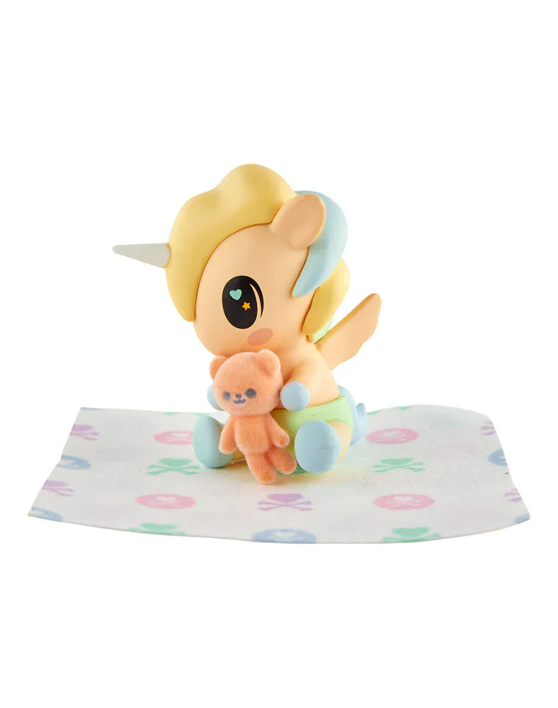 A tokidoki Baby Unicorno Blind Box figure with a yellow mane holds an orange teddy bear on a patterned cloth—a delightful collectible for fans of tokidoki.