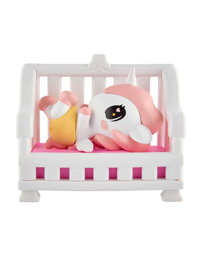 A tokidoki Baby Unicorno Blind Box figurine with a pink mane lies on its back on a white bench with a pink cushion, holding a yellow object—ideal for fans of collectible figures.