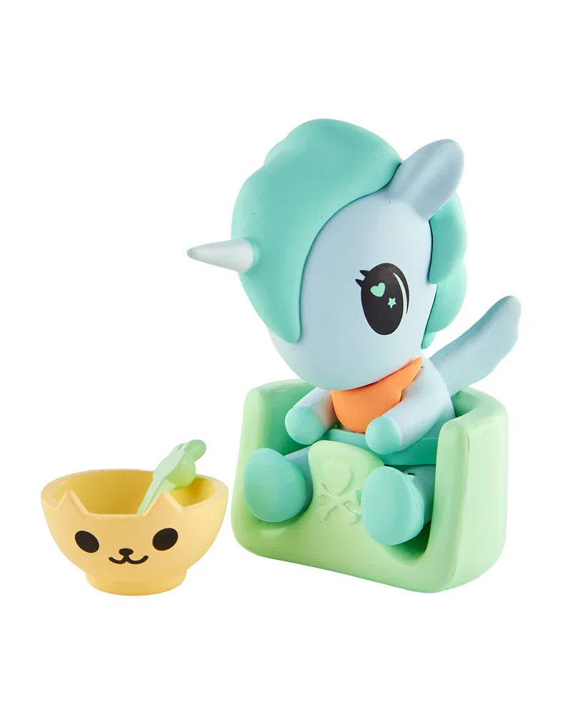 A tokidoki Baby Unicorno from the Tokidoki Baby Unicorno Blind Box sits on a green chair beside a yellow cat-face bowl and green spoon, perfect for tokidoki collectors.