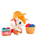 A tokidoki Farmers Market Unicorno Blind Box figure with a straw hat sits by a basket and berry cartons on a pink crate—an adorable collectible for any tokidoki fan.