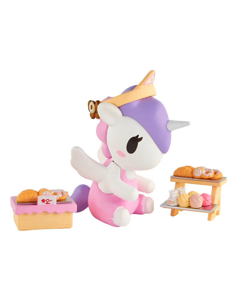 A pink and white tokidoki Farmers Market Unicorno Blind Box figure with a purple mane sits beside miniature bread and pastry displays—perfect for fans of collectible figures by tokidoki.