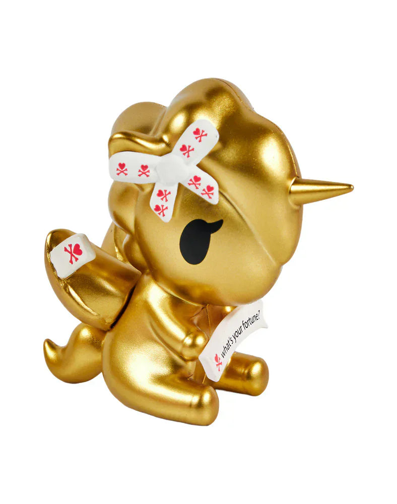 A charming Fortune Cookie Unicorno from Tokidoki sits with closed eyes, wearing a white bow with red symbols and holding a fortune slip—get this surprise figure in the Fortune Cookie Unicorno Blind Box!.
