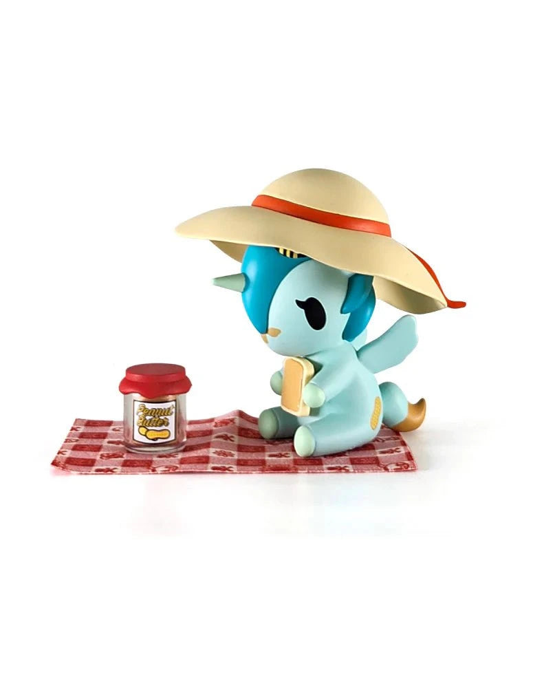 A small blue cartoon animal in a wide-brimmed hat sits on a red-checked picnic blanket, holding a sandwich by a 