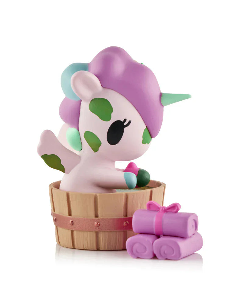 A cute Tokidoki Spa Day Unicorno Blind Box figurine with a purple mane and green spots sits in a wooden tub beside three rolled pink towels tied with ribbon, capturing the spa day charm of this collectible Unicorno toy.