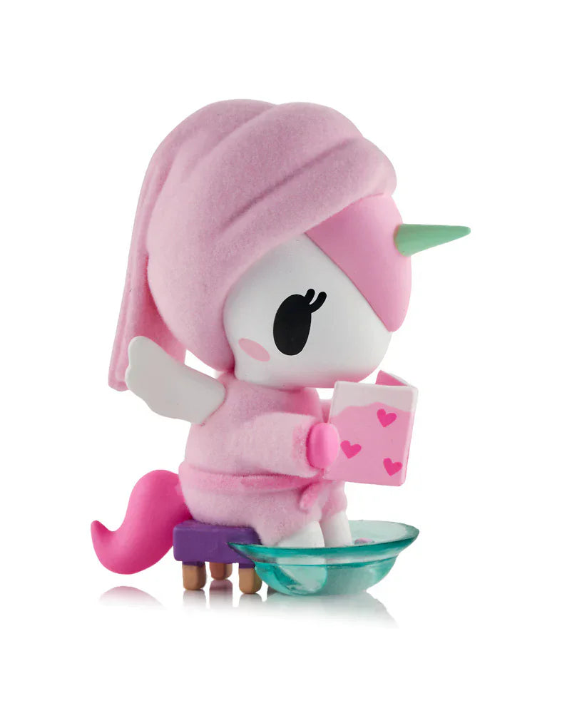 A Tokidoki Spa Day Unicorno Blind Box figurine with pink hair and a towel relaxes on a bench, holding a heart book and dipping its feet in a glass bowl for the ultimate kawaii wellness vibe.