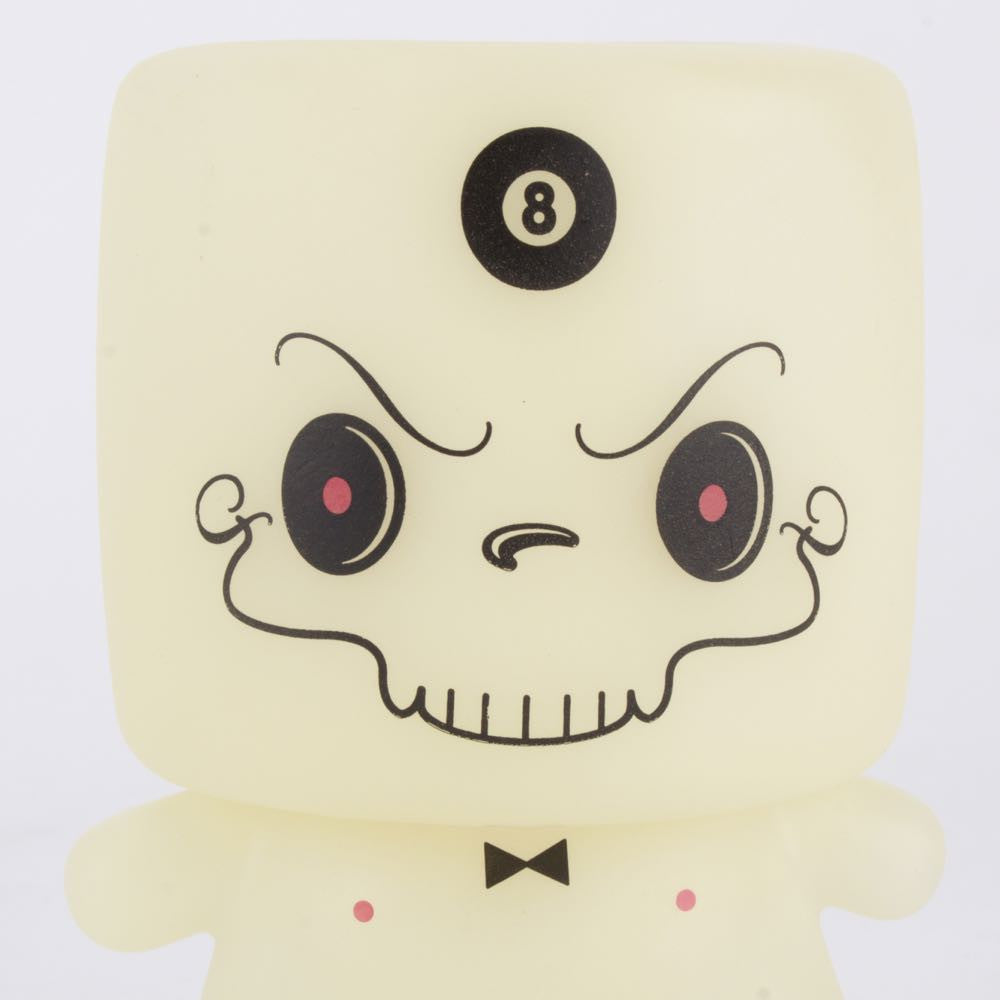 Introducing the limited edition figure, Grim Marshall by 64 Colors: a cream-colored toy with a square head displaying a black eight ball symbol. Its face boasts large black eyes with red centers, a curly mustache, a wide grin, and is adorned with a small black bow tie. Released by Squibbles Ink + Rotofugi (US), this character is sure to captivate collectors.