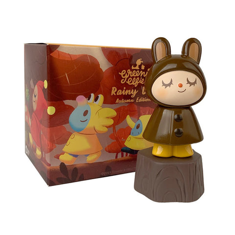 A Greenie & Elfie Rainy Day Autumn Edition Blind Box vinyl toy of a character in a brown raincoat styled like a bear, with a sleepy expression, displayed next to its colorful packaging box featuring autumn series artwork.